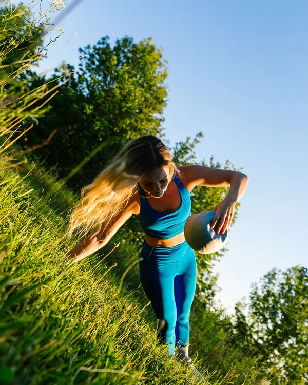 Woman in blue activewear carrying a Pilates ball down a grassy hillside